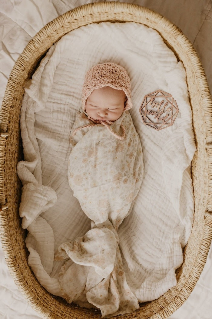 baby girl in her newborn announcement photo inside a baby basket wearing blush pink bonnet and wrapped in her meadow cream swaddl blanket made with natural cotton fabric