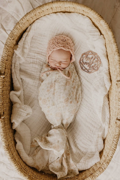 baby girl in her newborn announcement photo inside a baby basket wearing blush pink bonnet and wrapped in her meadow cream swaddl blanket made with natural cotton fabric