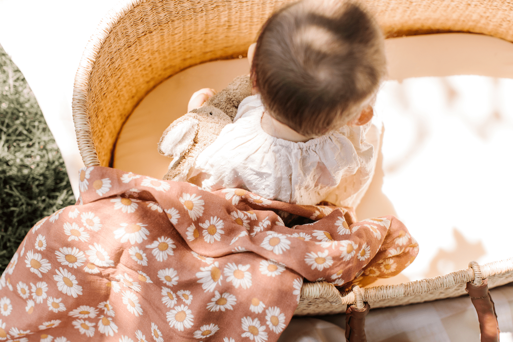 A baby in a basket holding a bunny toy, with a Mini Wander earthy clay brown color swaddle laid on the basket's side.