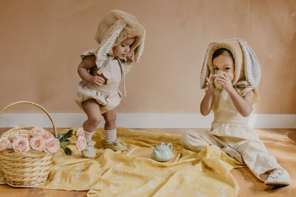 teaparty setup for two darling little girls in neutral baby clothes and animal hats sitting and standing on top of the yellow muslin blanket