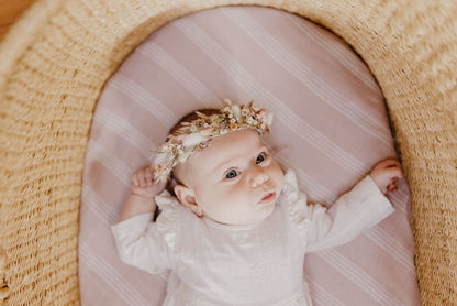 Little girl with a flower crown wearing white baby clothes laid down on her baby bassinet wrapped in a smoky rose mauve color receiving blankets