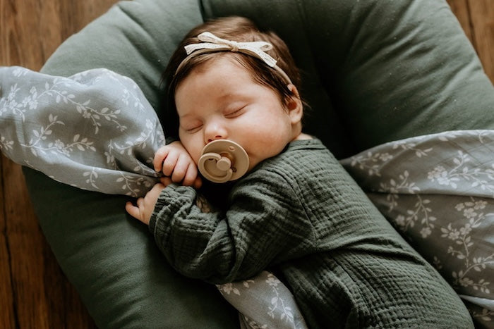 an adorable little baby wearing green boho baby romper, a lace ribbon headband comfortably lounging in her baby lounger using the bloom cotton blanket and her arms as the pillow while sleeping and sucking her taupe colored pacifier