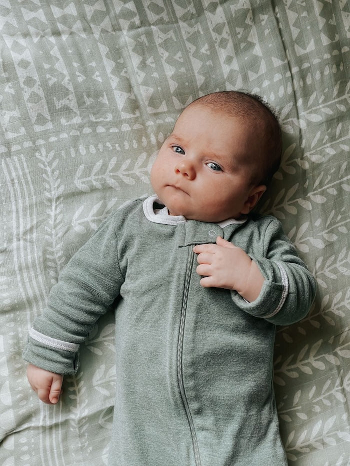 photo of a baby boy looking at the camera wearing a green onesie lying on our tapestry green spruce boy swaddle which is a perfect gift for baby