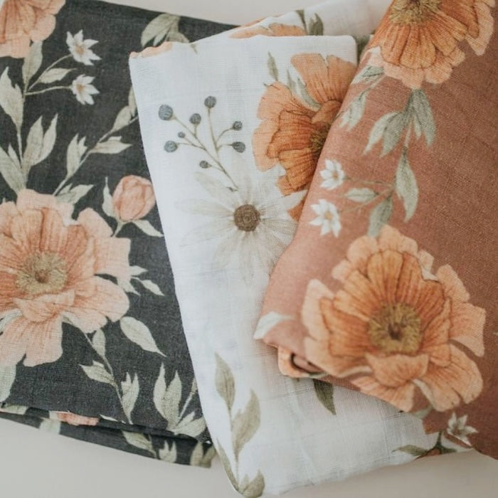 three folded cotton swaddle blankets side-by-side in different colors, gray, white and clay, showing the peony blooms in the peony swaddle