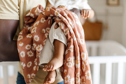 A baby is being carried by his father, with a Mini Wander earthy clay brown color with Daisy floral design swaddle draped over the baby's legs.