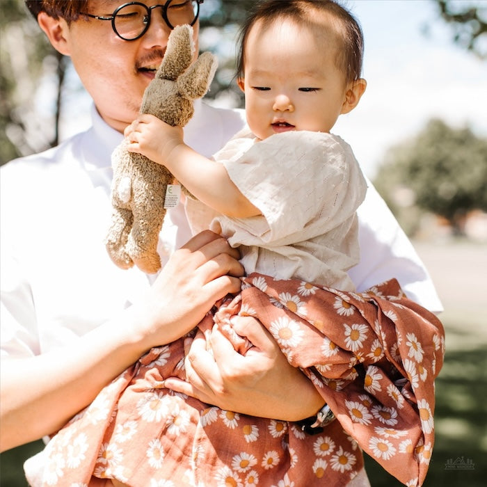 A baby is half wrapped in an earthy clay brown color with Daisy floral design swaddle while being carried by his father.