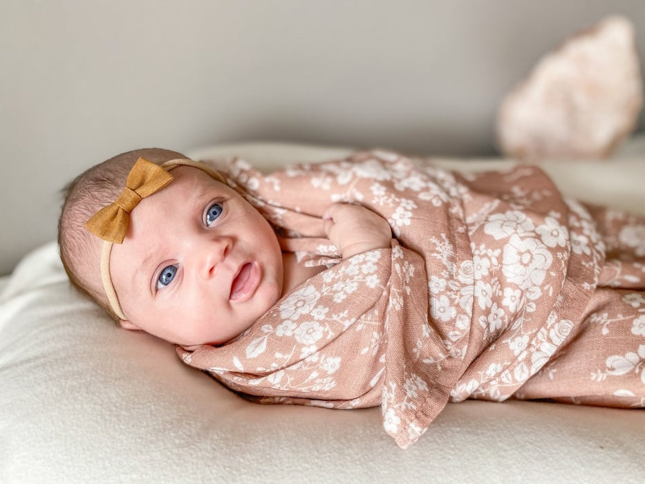 A baby wearing an amber-colored bow is wrapped in a sienna-colored swaddle with a white floral design