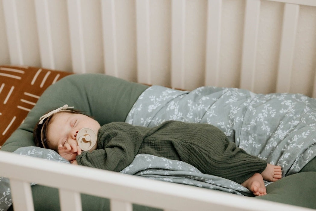 sweet baby with a baby pacifier sleeping peacefully in her white wooden crib with the floral muslin swaddle used as a bed cover