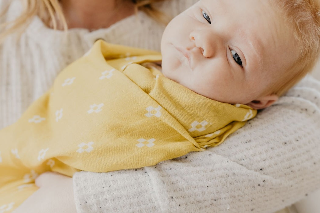 sleepy baby looking at the camera in a swaddling muslin blanket wrapped in yellow baby blanket