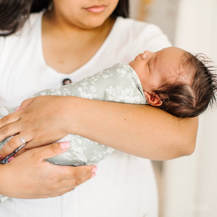 A mother holds her baby, who is tightly wrapped in a sage green swaddle.