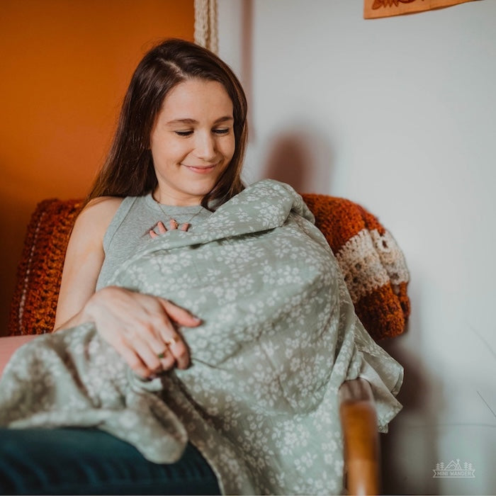 A mother sits and holds her child, who is wrapped loosely in a Mini Wander sage green swaddle.