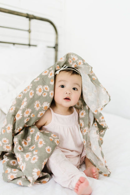 baby girl playing peek a boo with a lightweight muslin blanket. Baby blanket is a sage green color with lots of daisy flowers.