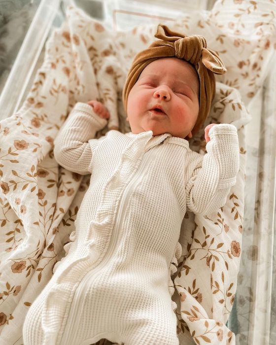 new baby girl dressed in a white ruffle waffle bodysuit with a brown bow, laying on a soft floral muslin baby blanket from Mini Wander