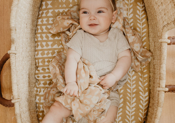 a baby girl laying in a moses basket that is lined with a boho geo patterned dark yellow cotton blanket