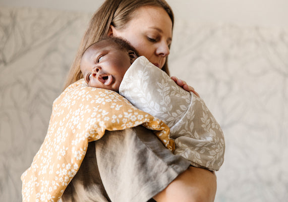 a mama holding her newborn son, wrapped in a neutral gray leaf swaddle blanket. a mustard yellow blanket is being used as a burp cloth