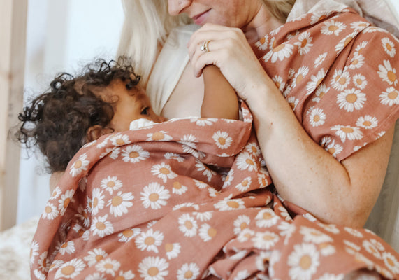 a mama is nursing her baby, she is using a red brown blanket as a breastfeeding cover. blanket has pretty daisies as a pattern