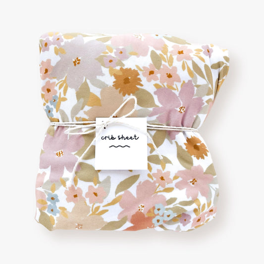 Jersey polyester crib sheet with Boho Jungle Floral pattern in lilac, beige, pink, orange, blue, and clay red flowers, sage green and yellow leaves, displayed on a crib mattress.