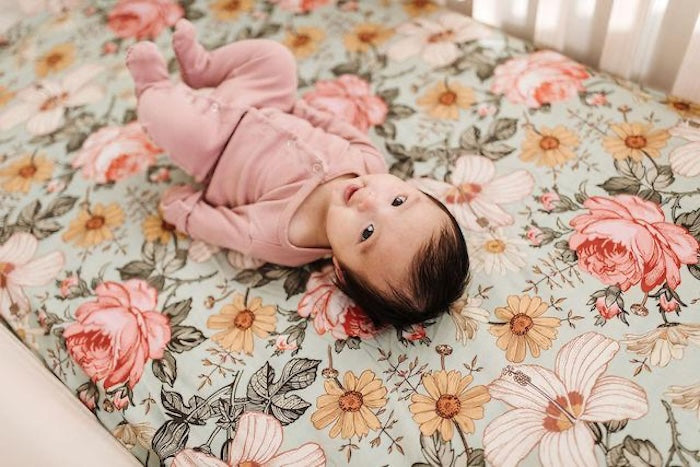A baby girl in a pink footie is lying down on a floral cribsheet with a vintage design that includes daisies, roses, and creamy pink hibiscus.