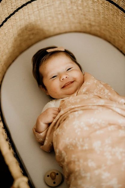 a happy baby smiling in her gummy smile lying on her baby bassinet wrapped in her peach muslin swaddle