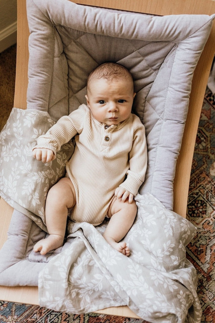 a baby boy sitting on a wooden lounger with a gray cover using the muslin swaddle blankets boy leafy sprig gray