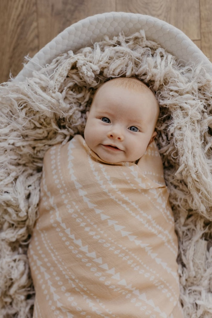 Smiling baby in lying in a baby basket with a textured throw swaddled in her tribal muslin swaddle wrap which is a peachy pink color.