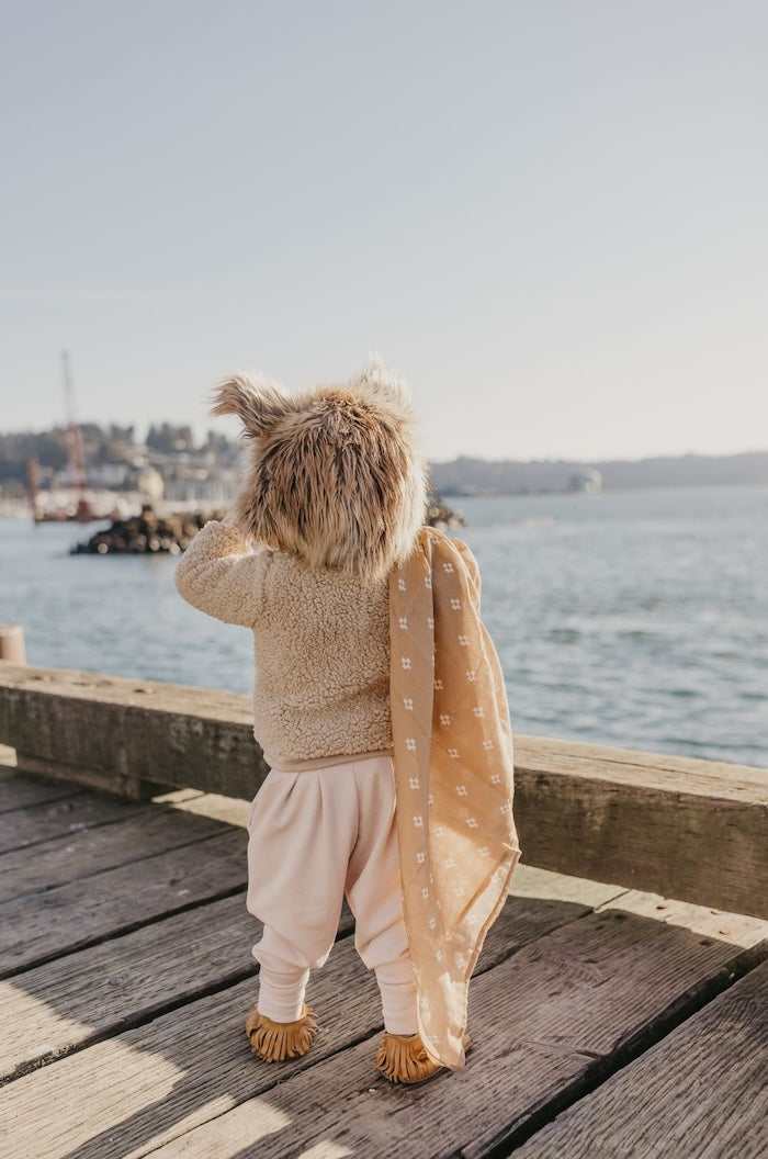 baby looking at the ocean wearing a neutral colored clothes and furry hat with a brown boho baby blanket on the shoulder