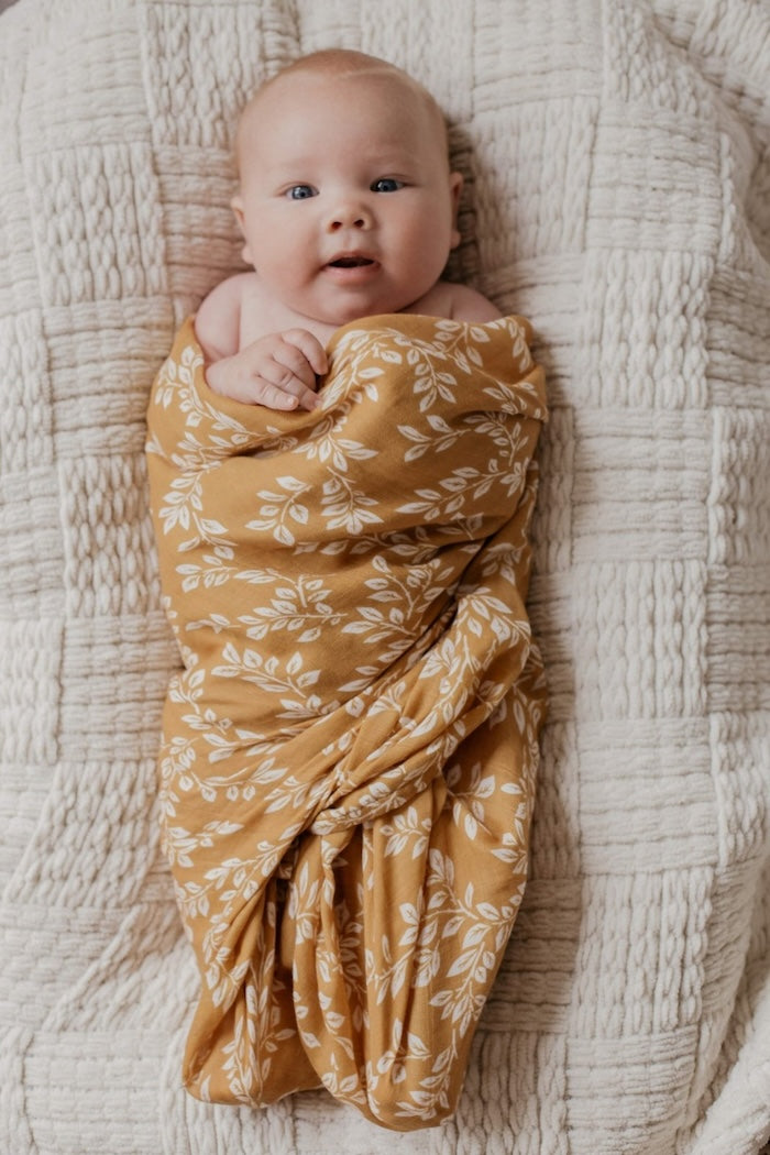 a happy baby looking towards the camera lounging comfortably in the baby lounger and baby is wrapped in the mustard leaf swaddle blanket