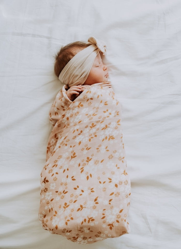A baby in a bandana is tightly wrapped in the Mini Wander peachy pink daisy swaddle blanket.