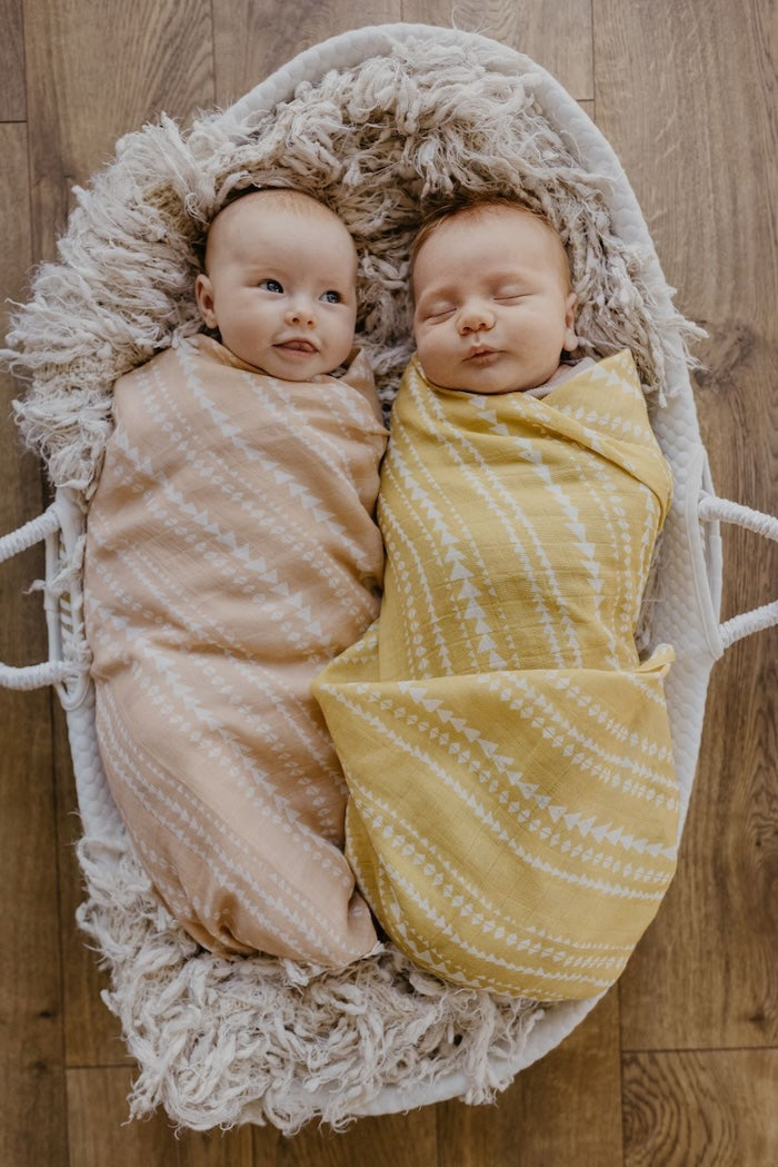 a beautiful image of two cute babies wrapped in our peach and dusty yellow aztec pattern nursery blankets laid down in a baby basket covered with a light brown faux fur