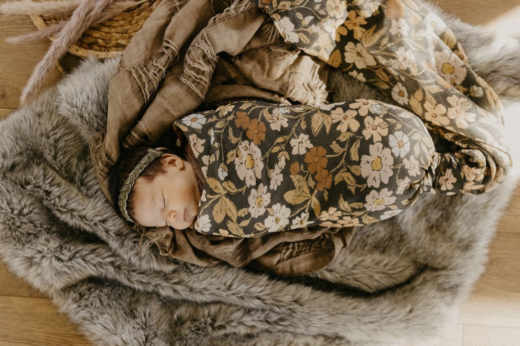 a dreamy photo of a little girl with metallic green leaves headband wrapped around in our thicket flower swaddle sleeping and laid down on a mix of textured fabric and faux fur
