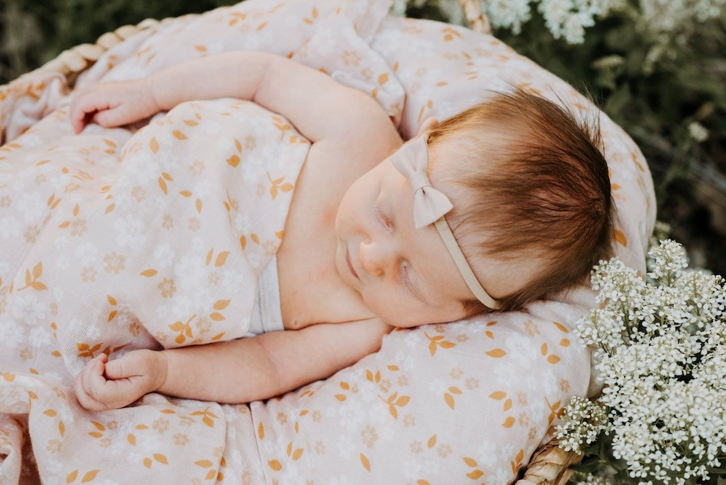 A pictorial viewed on the side of a sleeping baby in a basket with a pink bow tucked in and a peach floral daisy swaddle.