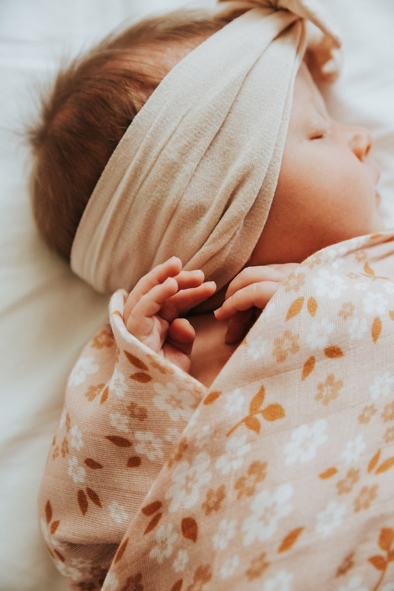 A close-up of a baby wearing a headwrap and sleeping on a peach-colored swaddle.