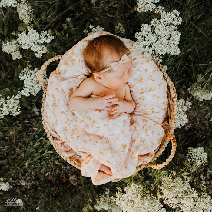 a newborn baby girl in a basket in the field with a tiny pink bow and a pink swaddle blanket with orange, beige and white flowers.