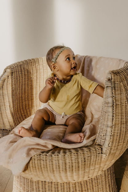 baby girl wearing yellow and earthtone baby clothes sitting and glancing on a rattan chair covered with pastel pink baby blanket