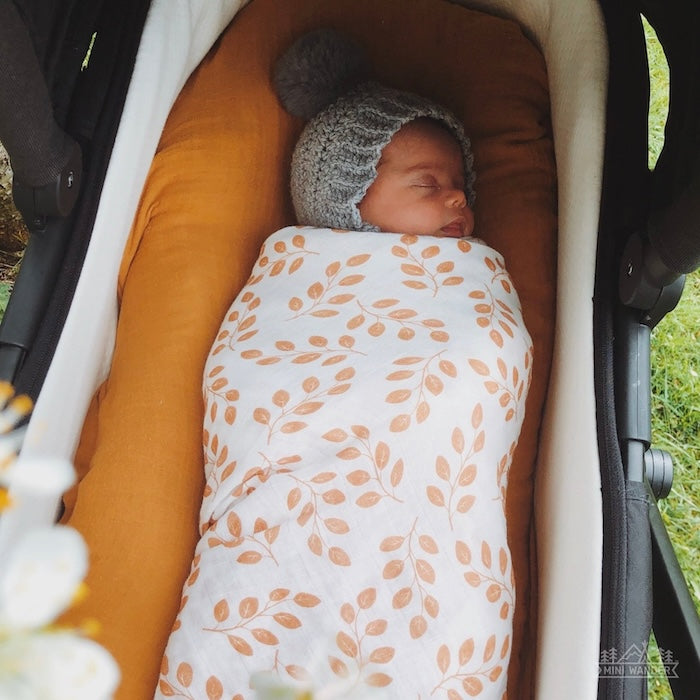 baby with a gray bonnet peacefully sleeping on her stroller and swaddled in her leaves amber swaddle blanket