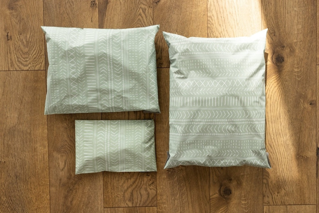 An image comparing different sized poly mailers. Pastel sage green color with white block printed boho line design. Pictured are sizes 12x15.5 , 10x13 and 6x9 self sealing plastic shipping envelopes.
