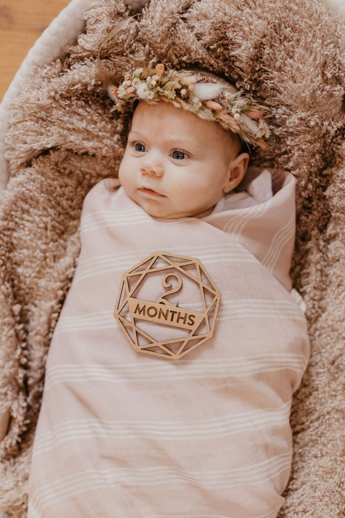Little baby girl with a flower crown in her 2 month baby milestone laid down on a furry surface and wrapped in our classic stripe blankets in the the smoky rose mauve color