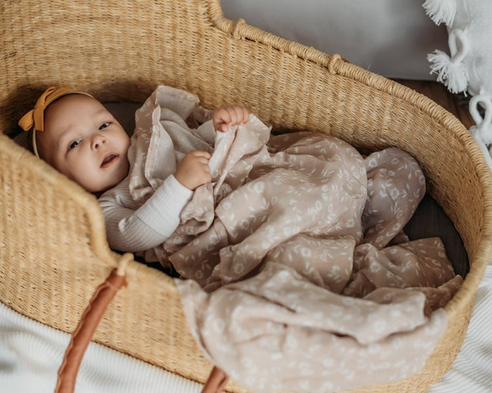 an adorable baby inside a baby basket holding her wildflowers taupe baby wrap in her hands while using it as a floral baby blanket