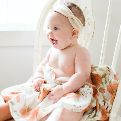 an adorable sitting and covered in our white peony swaddle, wearing a white headband while smiling