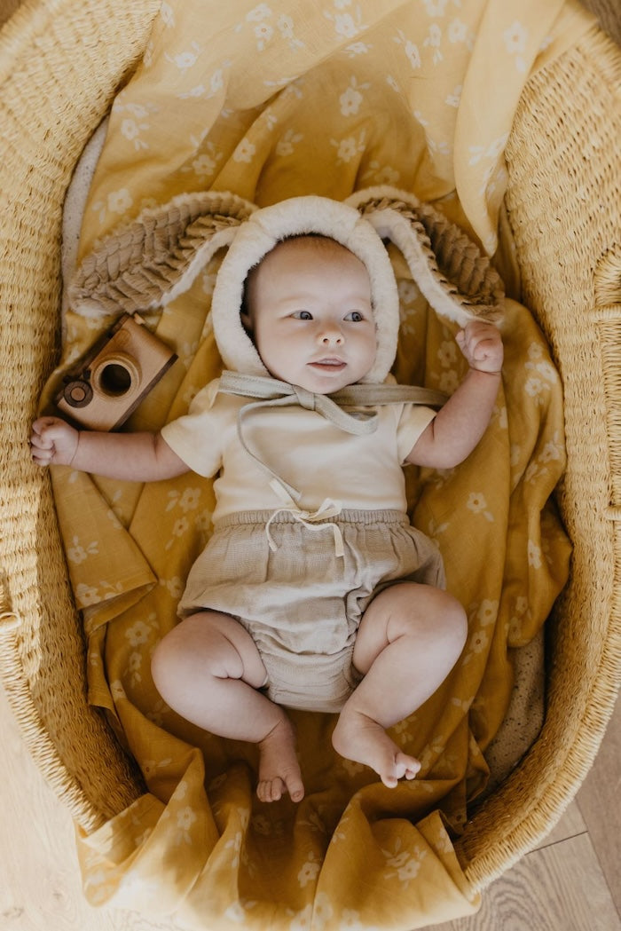 smiling toddler wearing neutral baby clothing and a furry animal hat inside the baby bassinet covered with yellow muslin blanket