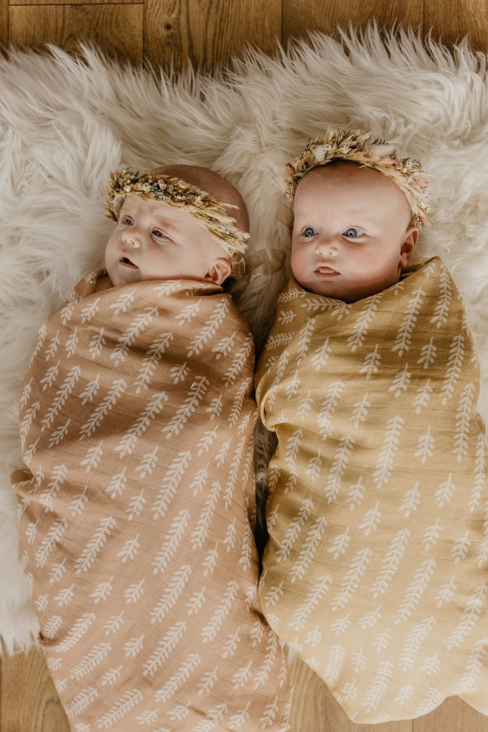 adorable twin babies wearing flower crowns and swaddled in the wheat swaddling blankets on top of white faux fur background looking perfect in their muslin blankets