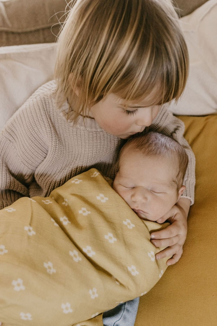 Big brother wearing neutral baby clothes kissing baby swaddled in a yellow baby blanket