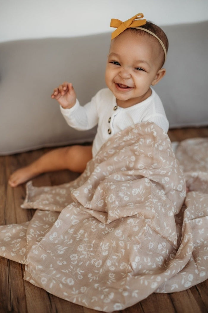 an adorable baby wearing wwhite baby clothes and mustard headband sitting on a wooden floor holding the wildflowers taupe baby wrap in her hands