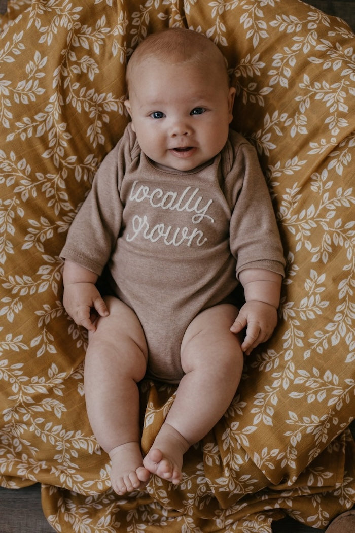 chubby and chunky baby wearing a brown body suit that says locally grown sitting comfortably in his chair draped and covered with leaf swaddle blanket in the mustard color