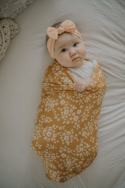 A baby in a pink bow is wrapped in a mustard baby swaddle wrap.
