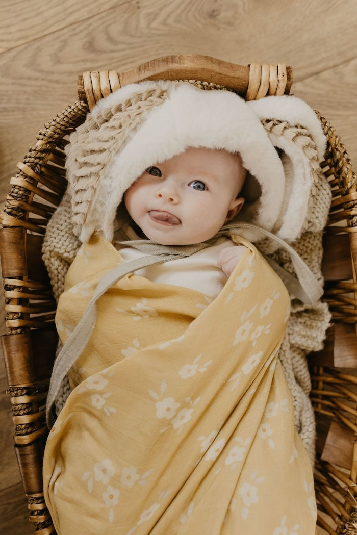 little baby wearing a furry hat tongue's out wearing the best seller yellow muslin blanket