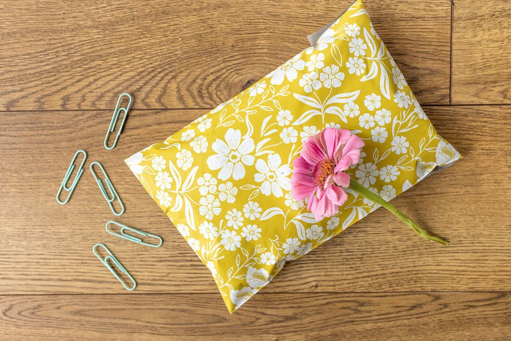 flatlay photo of the yellow 6x9 polymailer shipping bag laid flat on a wooden table with a pink flower on top. Beside it are five pieces of paper clips 