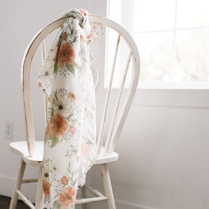 a glimpse of a boho nursery decor using The Mini Wander Spring Blossom floral baby swaddle tied in a rustic chair placed beside a window with white lace curtains