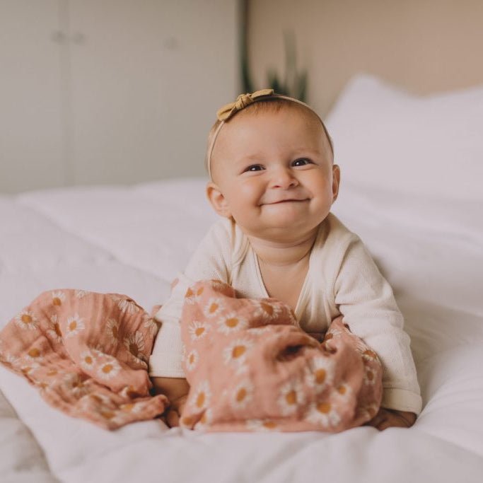 A baby with brown bow smiling with for someone in a crawl position and cuddled in a Mini Wander pinkish floral swaddle.