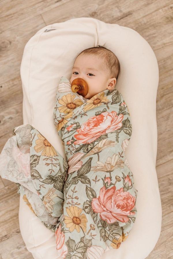 A swaddled baby with a pacifier, wrapped in a green swaddle with a floral design of roses, daisies, and hibiscus, one of Mini Wander's best-selling items.
